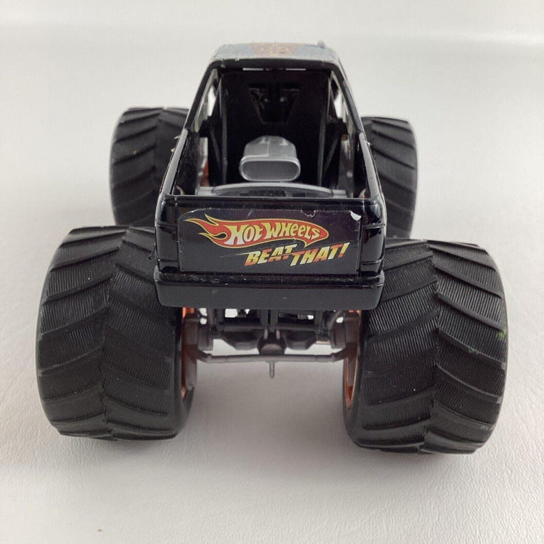 Puede incluir: Un juguete de cami&oacute;n monstruo Hot Wheels negro con neum&aacute;ticos negros grandes y detalles en naranja. El cami&oacute;n presenta el texto "HOT WHEELS BEAT THAT!" en la parte trasera. El juguete tiene un interior negro y un motor plateado.