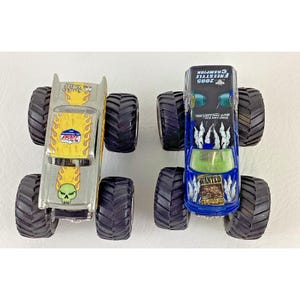 K&ouml;nnte beinhalten: Zwei Spielzeug-Monstertrucks. Einer ist silberfarben mit gelben Flammen und einer Totenkopf-Grafik. Der andere ist blau mit einer "WANTED"-Grafik und dem Text "2005 Freestyle Champion". Beide haben gro&szlig;e schwarze Reifen.