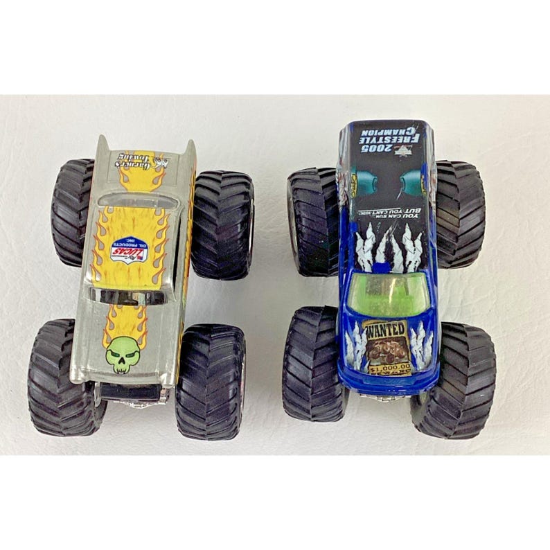 K&ouml;nnte beinhalten: Zwei Spielzeug-Monstertrucks. Einer ist silberfarben mit gelben Flammen und einer Totenkopf-Grafik. Der andere ist blau mit einer "WANTED"-Grafik und dem Text "2005 Freestyle Champion". Beide haben gro&szlig;e schwarze Reifen.