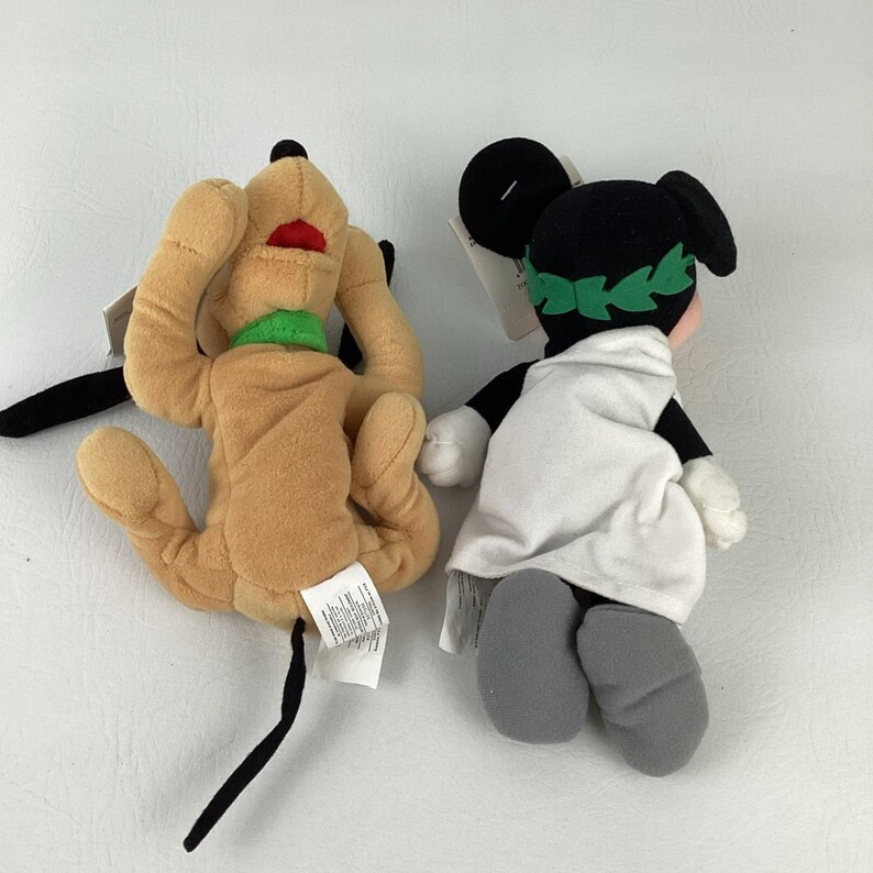 May include: Two plush toys featuring a tan dog with a green collar and a black-eared character wearing a white toga and a green leaf crown. Both toys are shown from the back, with white fabric tags attached to their bodies and limbs visible.