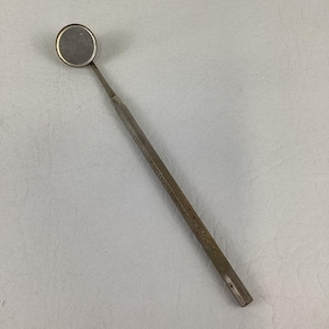 May include: A silver-colored metal dental mirror featuring a circular reflective head attached to a long, slender, hexagonal handle. The handle is engraved with the text MADE IN U.S.A. and the number 5, indicating its origin and specific model type.
