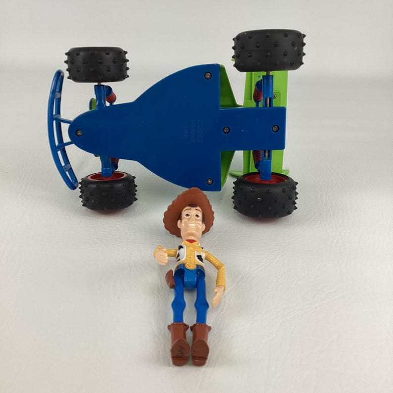 May include: A blue and green toy car with black tires and red accents, and a Woody action figure. The car is upside down, revealing its blue underside. The Woody figure is wearing a cowboy hat, yellow shirt, blue pants, and brown boots.