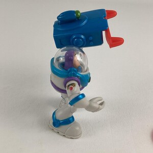 Op de afbeelding: Een Buzz Lightyear actiefiguur uit de Toy Story-franchise. De figuur is voornamelijk wit, met blauwe en paarse accenten. Het heeft een doorzichtige helm, een blauw jetpack en beweegbare ledematen. De figuur staat op een witte ondergrond.