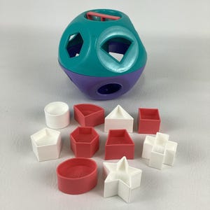Puede incluir: Un juguete clasificador de formas colorido con un recipiente en forma de bola de color verde azulado y púrpura. El recipiente tiene varios recortes geométricos. Se incluyen formas geométricas rojas, blancas y rojas, como un cilindro, un triángulo y una estrella.