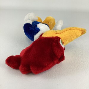 Op de afbeelding: Een pluche speelgoed van een tekenfilmvogel, met een grote rode kop, gele snavel en blauw-witte lichaam. Het oog van de vogel is zichtbaar en het speelgoed ligt op een wit oppervlak.