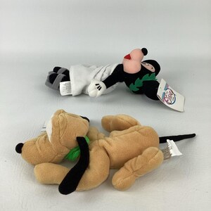 May include: Two plush toys featuring Disney characters. One is a black and white character wearing a white robe and a green wreath, with a Disney Store tag. The other is a tan dog character with black ears, a green collar, and a black tail.