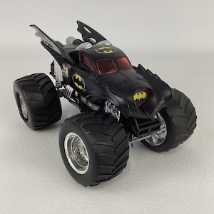 May include: A black monster truck toy with the Batman logo on the front, sides, and top. The toy has large, black tires and red tinted windows. The vehicle has a unique design with wing-like features and chrome accents.