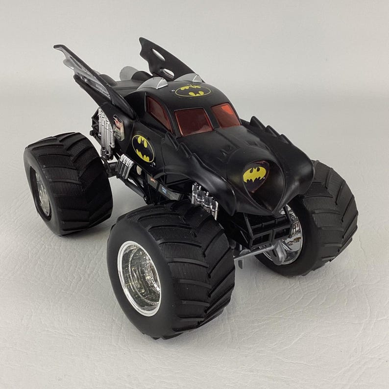 May include: A black monster truck toy with the Batman logo on the front, sides, and top. The toy has large, black tires and red tinted windows. The vehicle has a unique design with wing-like features and chrome accents.