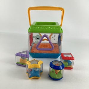 May include: A colourful baby toy set featuring a large, clear plastic cube with a green top and orange handle. The cube has a purple triangle and a mouse graphic. Smaller, clear plastic shapes with toys inside surround the cube.