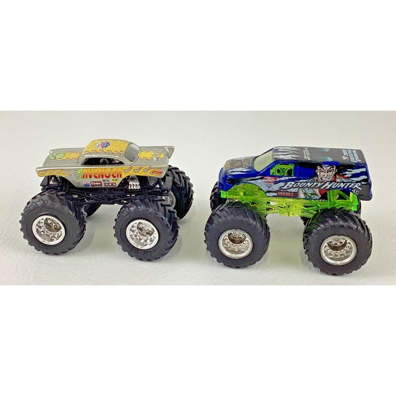 K&ouml;nnte beinhalten: Zwei Spielzeug-Monstertrucks. Einer ist silber und gelb mit dem Text "SIVENO CH", der andere ist blau und gr&uuml;n mit dem Text "BOUNTY HUNTER". Beide haben gro&szlig;e schwarze Reifen und detaillierte Designs.