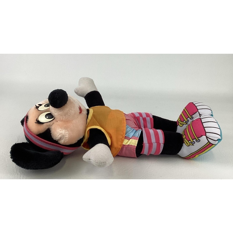 May include: A plush toy of a cartoon mouse character lying on its side. The character has black ears, a tan face, and a black nose. It wears an orange sleeveless top, pink and blue striped shorts, and white sneakers with pink laces and accents.