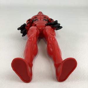 Puede incluir: Una figura de acci&oacute;n roja con detalles negros, vista desde abajo. La figura tiene una cabeza detallada con boca blanca y dientes afilados. Las piernas de la figura est&aacute;n separadas y sus pies son planos.