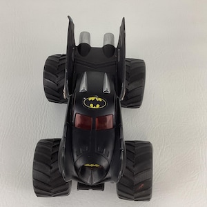 May include: Black toy monster truck with large, textured tires. The truck features a Batman logo and red tinted windows. The vehicle has two silver exhaust pipes and a bat-shaped design on the back.
