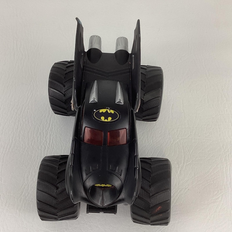 May include: Black toy monster truck with large, textured tires. The truck features a Batman logo and red tinted windows. The vehicle has two silver exhaust pipes and a bat-shaped design on the back.
