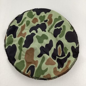 May include: Round camouflage pattern fabric in shades of green, brown, and black. The fabric has a black stitched edge. The pattern is reminiscent of military or hunting apparel.