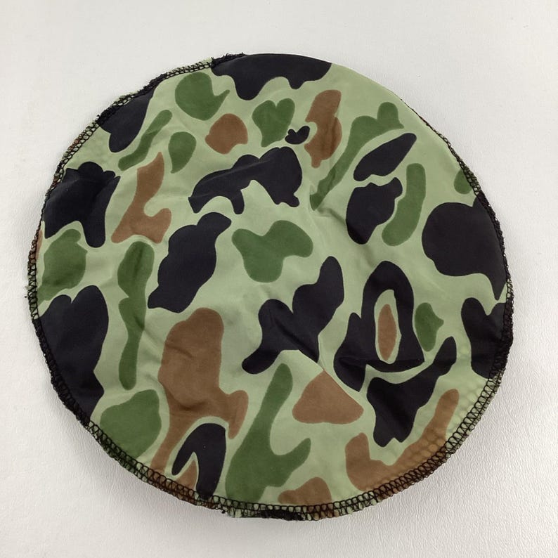 May include: Round camouflage pattern fabric in shades of green, brown, and black. The fabric has a black stitched edge. The pattern is reminiscent of military or hunting apparel.