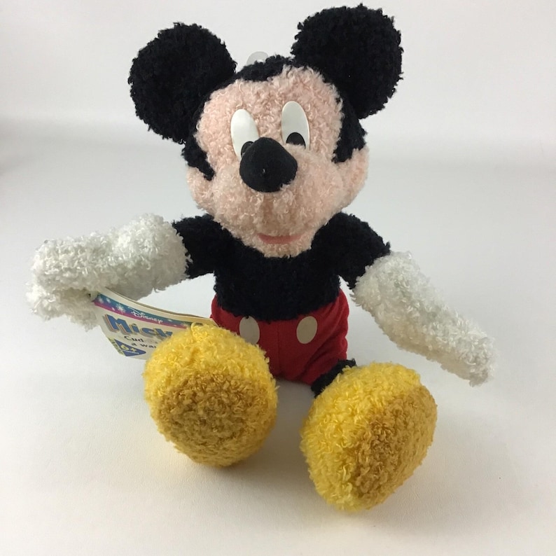 May include: A plush Mickey Mouse toy with black ears, a black body, red shorts, and yellow feet. The toy has white gloves and a tag attached. The face is pink with white eyes and a black nose.