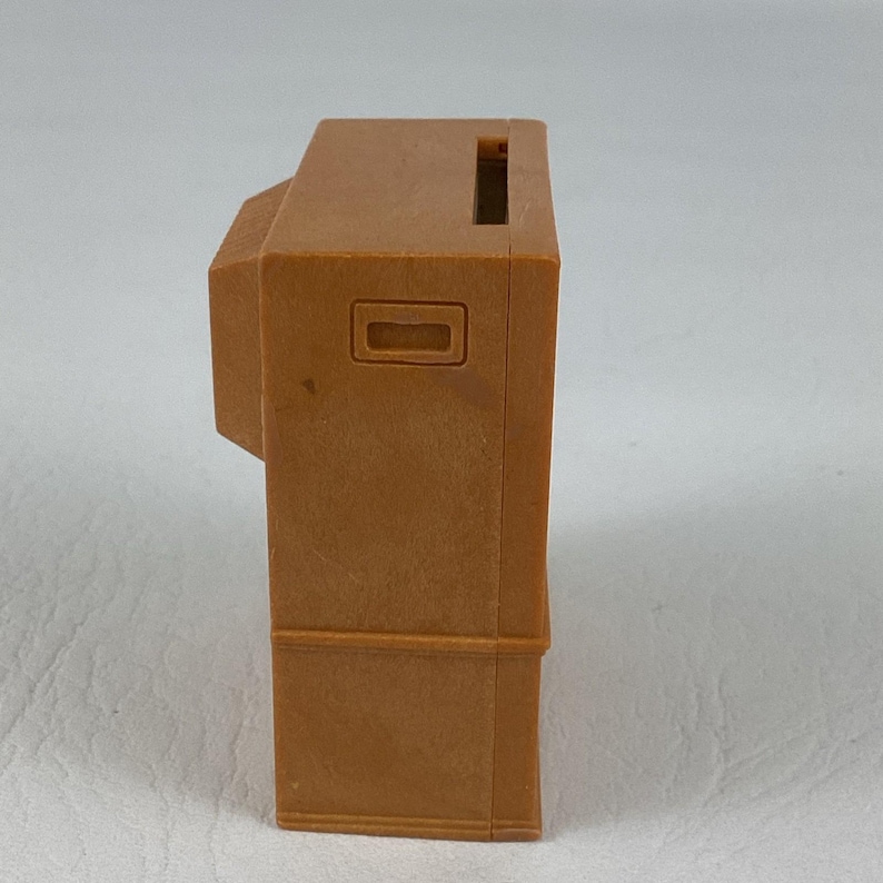 Op de afbeelding: Een bruine, rechthoekige speelgoedkassa van plastic, met een sleuf aan de bovenkant en een rechthoekige knop. Het heeft een klein, aangehecht gedeelte aan de linkerkant. Het speelgoed is bruin.