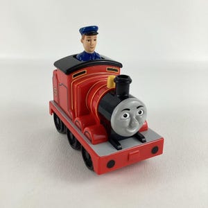 May include: A red toy train with black wheels and a smiling face. The train has a black top and a small figure of a person wearing a blue hat. The number 5 is visible on the side of the train.