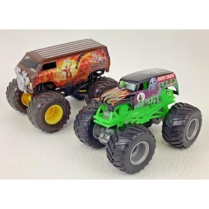 May include: Two toy monster trucks. One is a brown van with a graphic of a warrior and lightning. The other is a green and black truck with "Grave Digger" text and a skull graphic. Both have large black tires.