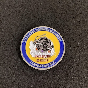 May include: A silver challenge coin with a blue and yellow border. The center features a black bull charging through a white cloud with the text "910th Civil Engineer Squadron" and "Prime Beef Leading the Way".