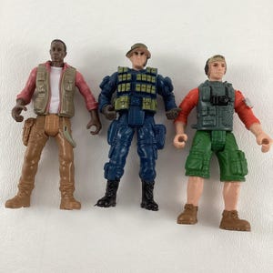 May include: Three action figures, one wearing a brown vest and pants, one wearing a blue and green military uniform, and one wearing a red and green vest and shorts.