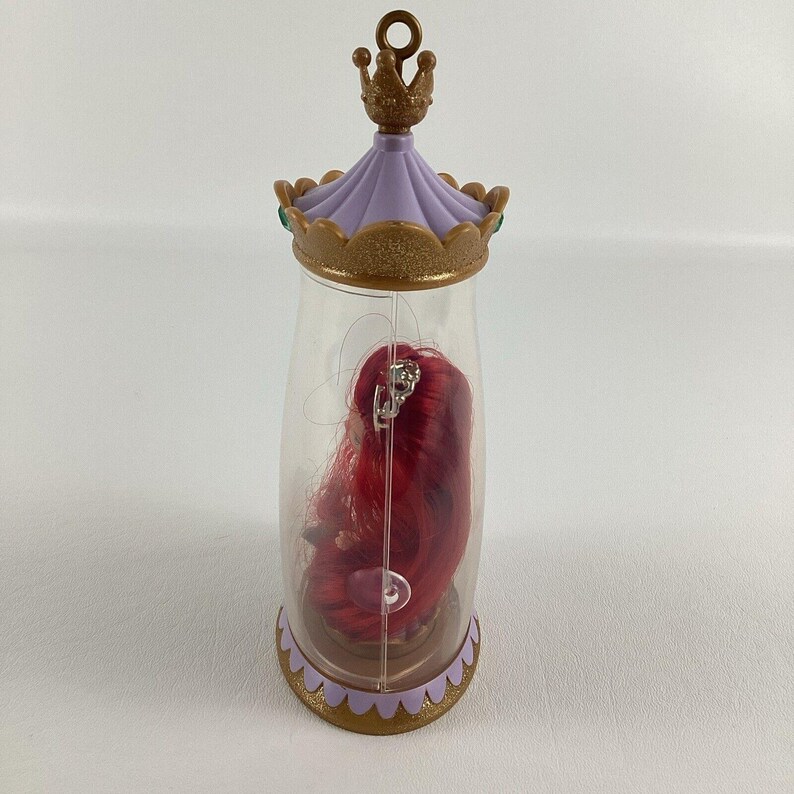 Puede incluir: Un recipiente de pl&aacute;stico transparente con una tapa y base dorada y lila en forma de corona. En el interior, se ve una peluca roja con una tiara plateada. El recipiente tiene un lazo en la parte superior para colgar.