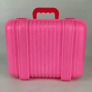 Puede incluir: Una maleta de plástico rosa brillante con un asa roja. La maleta tiene forma rectangular con esquinas redondeadas y una superficie acanalada. El asa es arqueada y está sujeta de forma segura en la parte superior.