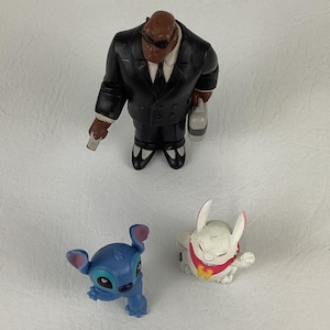 May include: A collection of Disney character figurines. The figures include a black-suited character with sunglasses, a blue alien-like creature, and a white rabbit-like figure with a red collar.