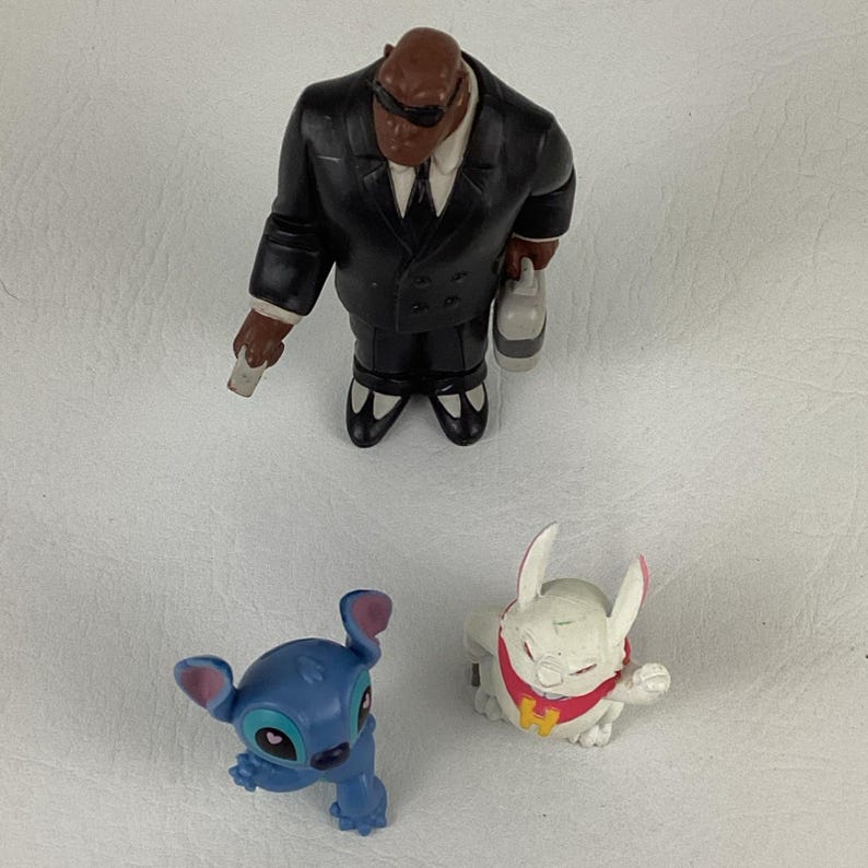 May include: A collection of Disney character figurines. The figures include a black-suited character with sunglasses, a blue alien-like creature, and a white rabbit-like figure with a red collar.