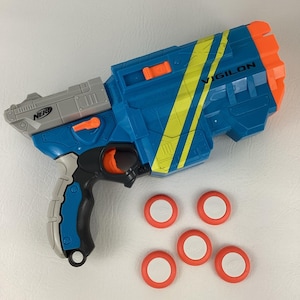 May include: A blue and orange Nerf Vigilón toy gun with yellow stripes and the word "VIGILON" printed on the side. Includes five red and white disc-shaped projectiles. The gun has a gray handle and trigger.