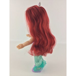 May include: A red-haired doll wearing a green and blue dress with seashells and a purple crown. The doll is wearing purple shoes.