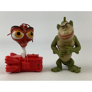 Può includere: Un veicolo giocattolo in plastica rossa con una grande testa stilizzata dotata di occhi gialli sovradimensionati e una parte superiore marrone, accanto a una statuetta di creatura umanoide verde e ruvida con petto beige e ampio sorriso.