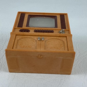 Op de afbeelding: Een miniatuurtelevisie in vintage-stijl in bruintinten. De speelgoed-tv heeft een scherm, bedieningsknoppen en kastdeuren met bloemendetails. Het bovenste gedeelte heeft een donkerbruin luidsprekerrooster.