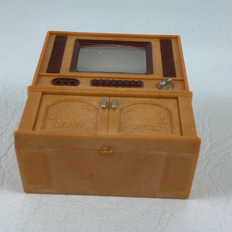 Op de afbeelding: Een miniatuurtelevisie in vintage-stijl in bruintinten. De speelgoed-tv heeft een scherm, bedieningsknoppen en kastdeuren met bloemendetails. Het bovenste gedeelte heeft een donkerbruin luidsprekerrooster.