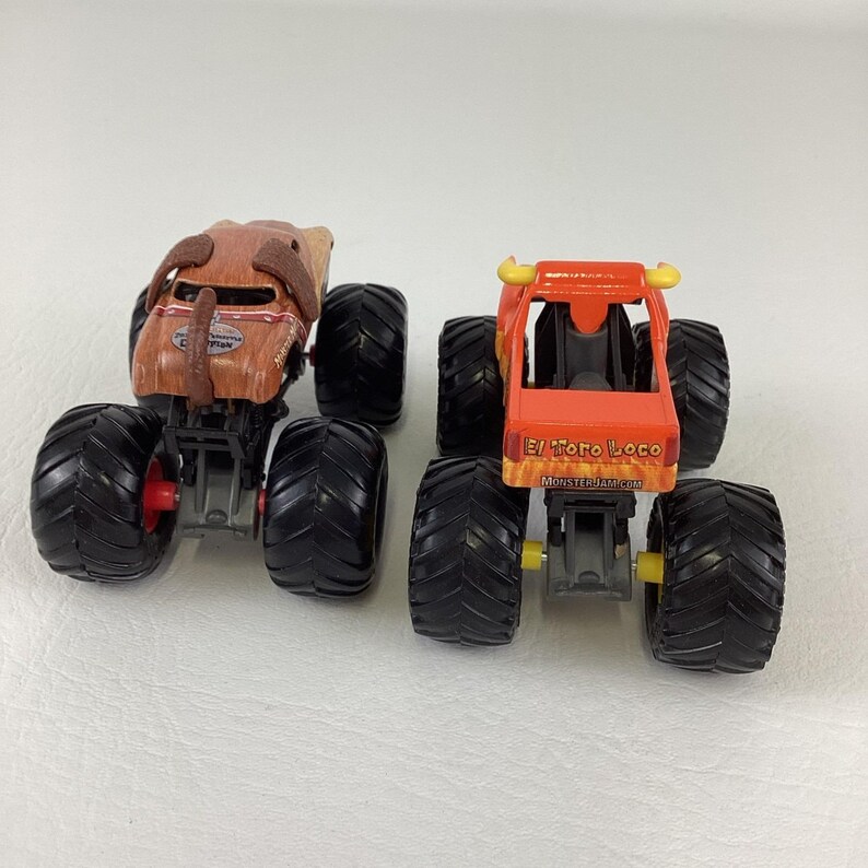 May include: Two toy monster trucks. One is brown with a dog-like design, the other is orange with yellow horns and the text "El Toro Loco". Both have large black tyres with red and yellow accents.