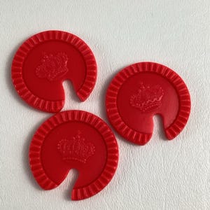 Op de afbeelding: Drie rode, cirkelvormige spelstukken met een kroonontwerp en een gebogen uitsparing. De stukken hebben een geribbelde rand en zijn gerangschikt op een wit oppervlak. De kroon is in het midden van elk stuk in reli&euml;f aangebracht.