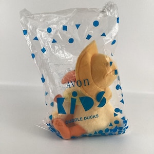 Puede incluir: Un pato de peluche amarillo con un sombrero amarillo con las palabras "Avon Kids Cuddle Ducks" impresas en el embalaje.