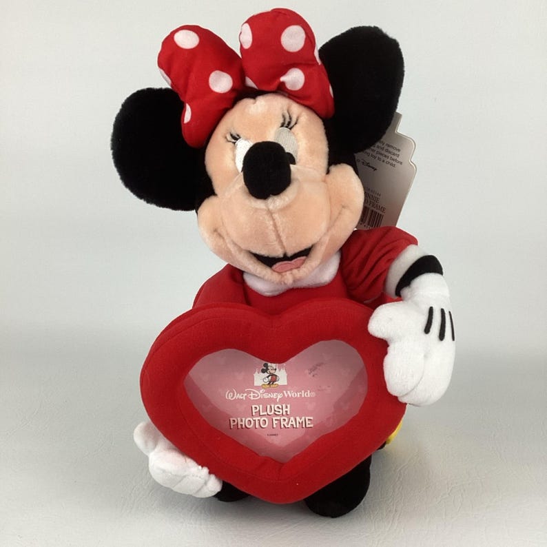 Pu&ograve; includere: Un peluche di Minnie Mouse con un vestito rosso e un fiocco a pois bianchi. Tiene in mano una cornice portafoto rossa a forma di cuore con la scritta "Walt Disney World Plush Photo Frame".