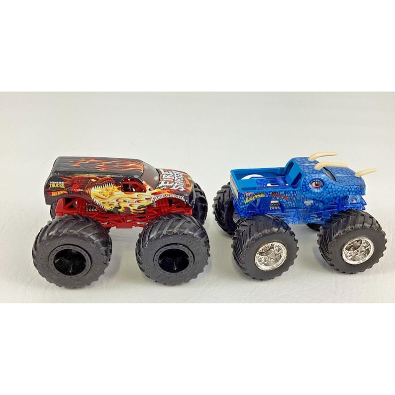 May include: Two toy monster trucks. One is red and black with a flame design and the text "Fire Storm". The other is blue with a dragon design. Both have large black tires.