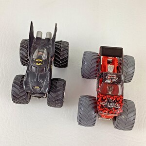 May include: Two toy monster trucks. One is black with a Batman logo, the other is red with a black and red pattern and the words "Northern Nightmare" and "2012 Freestyle Champion". Both have large gray tires.