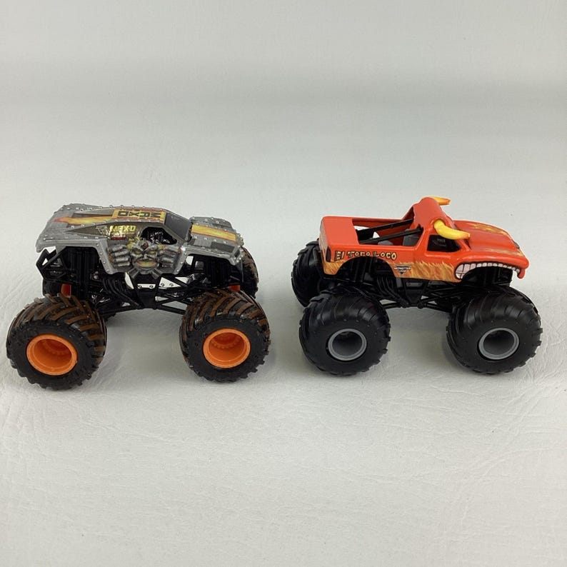 Puede incluir: Dos monster trucks de juguete. Uno es plateado y negro con ruedas naranjas, el otro es naranja con un dise&ntilde;o de toro y ruedas negras. Ambos tienen neum&aacute;ticos grandes y est&aacute;n dise&ntilde;ados para el juego todoterreno.