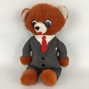 May include: A vintage plush teddy bear wearing a black and white pinstripe suit with a red tie. The bear has large, expressive eyes, a white muzzle, and orange fur on its head, paws, and ears.