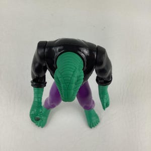 Puede incluir: Una figura de acci&oacute;n de pl&aacute;stico con una cabeza verde texturizada similar a la de un reptil, un torso negro y pantalones morados. La figura tiene brazos y pies verdes, con la cabeza inclinada hacia abajo. El dise&ntilde;o muestra una criatura humanoide.
