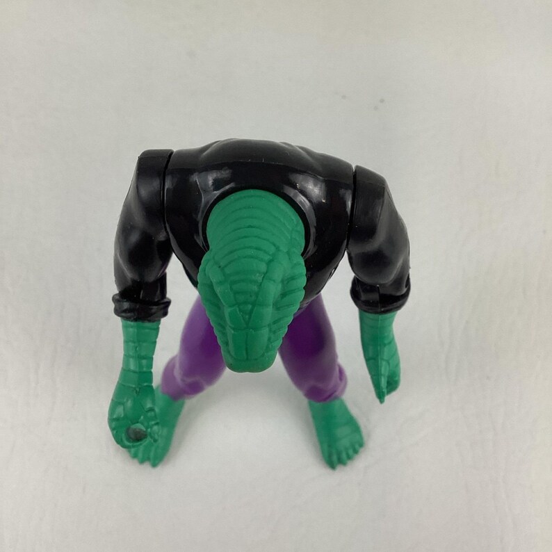 Puede incluir: Una figura de acci&oacute;n de pl&aacute;stico con una cabeza verde texturizada similar a la de un reptil, un torso negro y pantalones morados. La figura tiene brazos y pies verdes, con la cabeza inclinada hacia abajo. El dise&ntilde;o muestra una criatura humanoide.