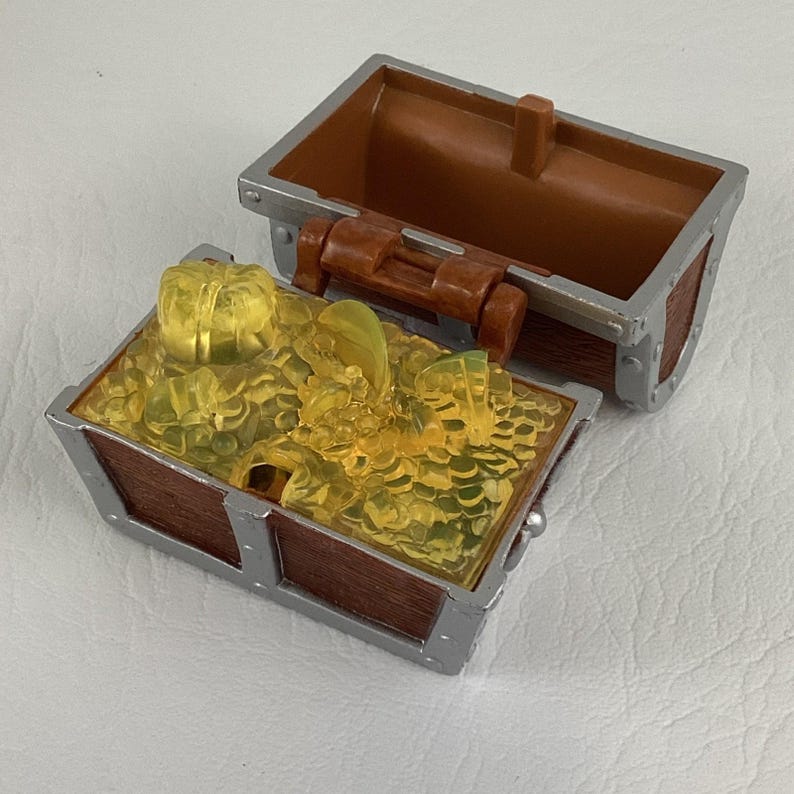 May include: A toy treasure chest, open to reveal a pile of translucent yellow gems. The chest is brown with silver metal accents and a brown lid. The lid is open, showing the inside of the chest.