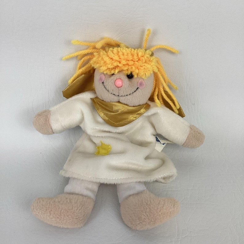 Puede incluir: Mu&ntilde;eca de peluche con una cara sonriente, cabello de hilo amarillo y un vestido blanco. La mu&ntilde;eca tiene una bufanda dorada y una peque&ntilde;a estrella amarilla en el vestido. Los brazos, las piernas y los pies de la mu&ntilde;eca son de color beige claro.