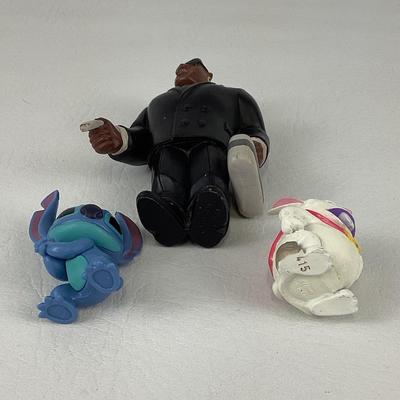 May include: Three small figurines: a blue alien-like character, a white rabbit with a pink hat, and a man in a black suit. The figures are made of plastic and are on a white surface.