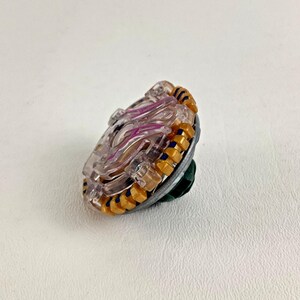 Puede incluir: Un trompo de juguete transparente y morado con detalles dorados y azules. El juguete tiene una base plateada y una punta verde. El trompo est&aacute; dise&ntilde;ado con m&uacute;ltiples capas y un anillo exterior texturizado.