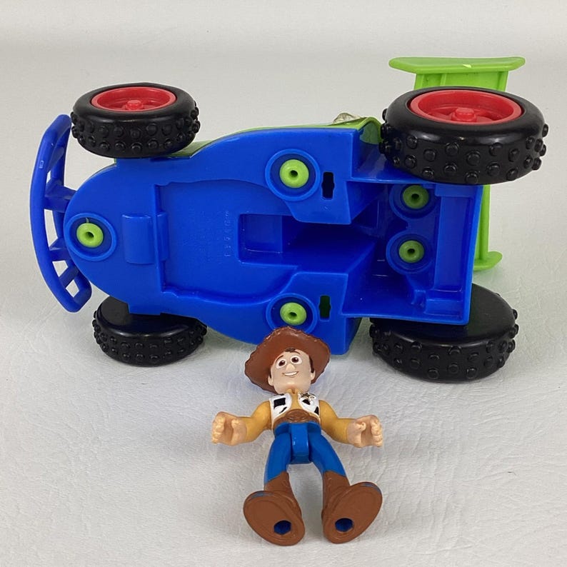May include: A blue toy car with black tires and red rims is flipped over, revealing its underside. A small Woody figure in a cowboy hat, vest, and blue pants is positioned in front of the car. The toy car has a green section at the back.